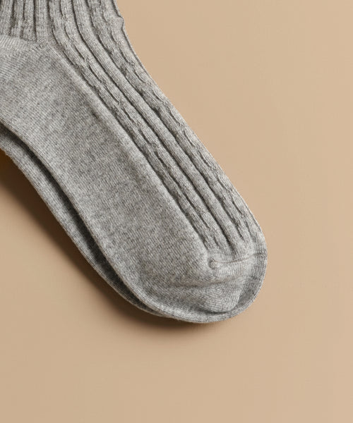 A close-up of Cable Socks in grey with a ribbed, textured knit pattern, ideal for everyday wear, shown lying flat on a beige surface.