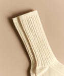 A pair of Cable Socks in cream lies flat on a beige surface, showcasing their ribbed knit cuffs overlapping slightly—ideal for everyday wear.