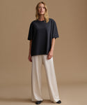 A woman wearing an oversized short-sleeve crewneck t-shirt in black paired with cream-colored pants, standing against a solid warm beige background.