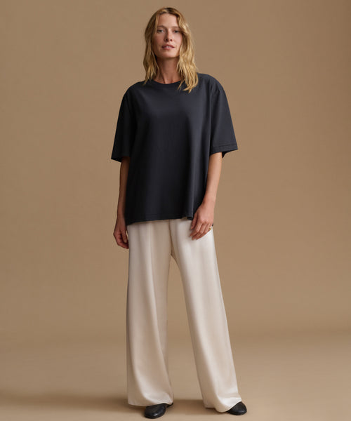 A woman wearing an oversized short-sleeve crewneck t-shirt in black paired with cream-colored pants, standing against a solid warm beige background.
