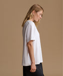 A woman in profile wearing a loose white short-sleeve t-shirt and black pants, standing against a solid beige studio background.