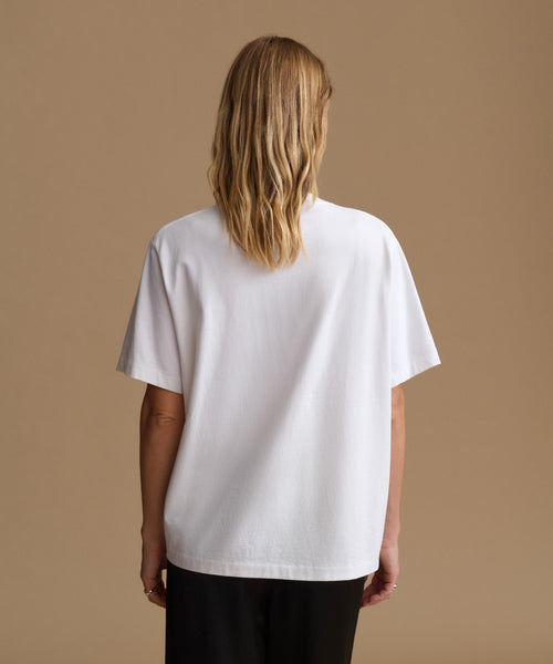 A woman viewed from the back wearing an oversized white short-sleeve T-shirt and black pants, standing against a plain warm beige studio background.