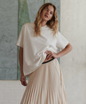A woman modeling an oversized off-white short-sleeve top and a pleated light beige skirt, standing against a textured light gray wall.