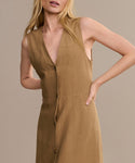 A woman wears the Callan Dress, a sleeveless tan vest dress with a deep V-neckline, standing against a beige background with one hand on her hip. Her face is partially out of frame. This classic piece is a timeless wardrobe staple.
