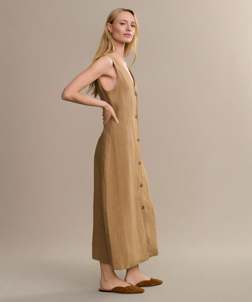 A woman stands sideways in the sleeveless, tan Callan Dress—a button-down midi with side pockets. She has long blonde hair, wears brown flats, and is set against a plain neutral background. A timeless closet staple.