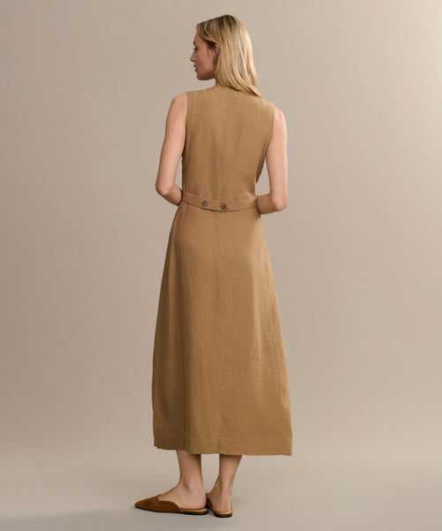 A woman with blonde hair stands facing away in the Callan Dress—a sleeveless, light brown vest dress with waist buttons—paired with flat brown shoes, set against a plain beige background.