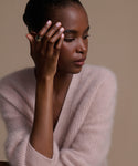 A woman in a pale pink fuzzy sweater rests her head on her hand, thoughtfully gazing downward. She wears the striking Campbell Cigar Band on her finger, and her short hair is neatly styled against a soft beige background.