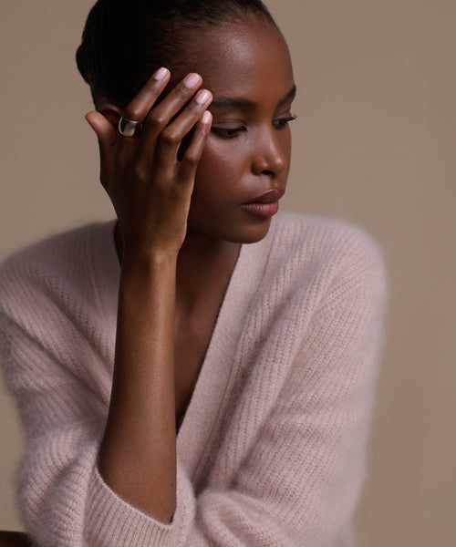 A woman in a pale pink fuzzy sweater rests her head on her hand, thoughtfully gazing downward. She wears the striking Campbell Cigar Band on her finger, and her short hair is neatly styled against a soft beige background.