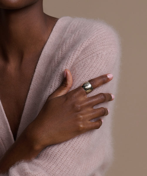 A person with medium-dark skin crosses their arms against a neutral beige background, wearing a soft pale pink fuzzy sweater and the chunky Campbell Cigar Band on their left hand.