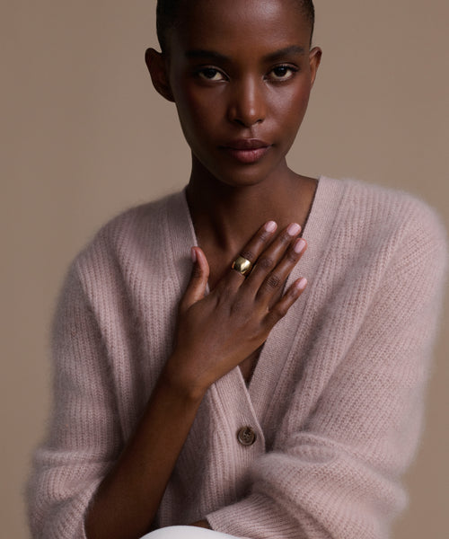 A woman in a soft, light pink cardigan poses against a beige background, resting one hand with the Campbell Cigar Band on her chest. She looks calmly at the camera with a neutral expression.