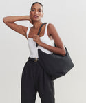 A woman in a white tank top and high-waisted black pants poses with one hand behind her neck, carrying the stylish Canvas Day Bag over her shoulder against a plain light background.