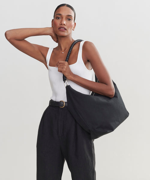 A woman in a white tank top and high-waisted black pants poses with one hand behind her neck, carrying the stylish Canvas Day Bag over her shoulder against a plain light background.