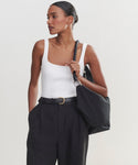 A woman wears a white sleeveless ribbed top, high-waisted black trousers with a belt, and carries a Canvas Day Bag over her shoulder. She stands against a plain light background, looking to the side.