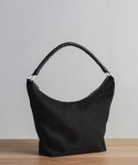 A black Canvas Day Bag with a twisted handle rests on a wooden surface against a light gray background. The bag features a curved zippered opening and showcases a minimalist, modern design.