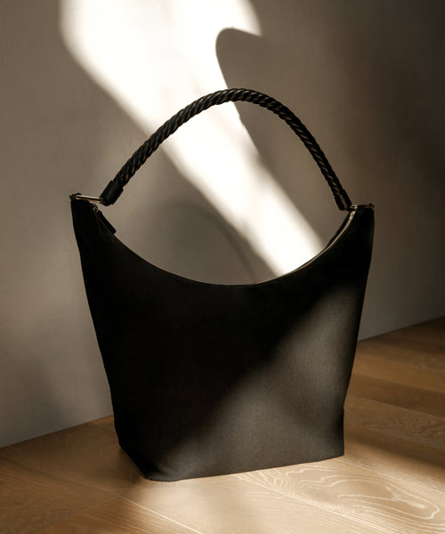 A Canvas Day Bag in classic black with a curved top and thick, twisted handle rests on a wooden floor, bathed in soft natural light and shadow.