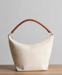 The Canvas Day Bag, featuring a cream-colored canvas body and a brown rope leather handle, is displayed on a wooden surface with a plain light gray background.