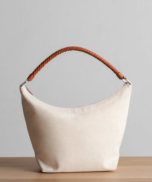 The Canvas Day Bag, featuring a cream-colored canvas body and a brown rope leather handle, is displayed on a wooden surface with a plain light gray background.