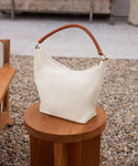 The Canvas Day Bag, featuring a white canvas body and a brown rope leather handle, is displayed on a round wooden stool amid outdoor furniture and gravel.