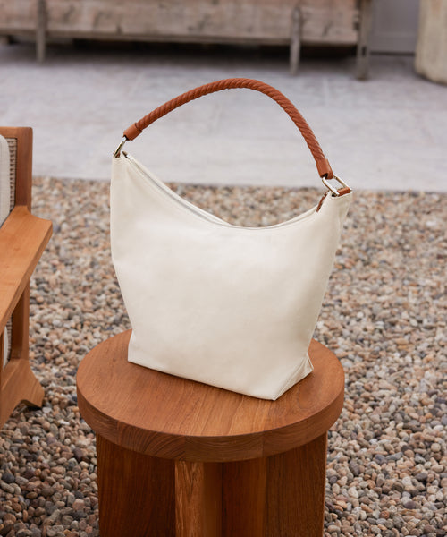 The Canvas Day Bag, featuring a white canvas body and a brown rope leather handle, is displayed on a round wooden stool amid outdoor furniture and gravel.