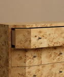 Close-up of a burlwood dresser with three drawers, featuring a light tan burl wood pattern with dark brown grain and small round dark metal knobs.