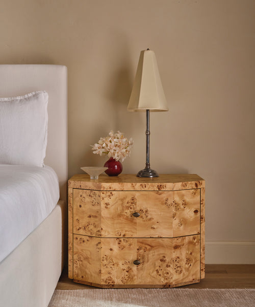 A two-drawer nightstand made of natural burl wood, styled in a bedroom next to a bed with white linens, holding a table lamp and a small vase of flowers.