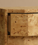 A close up of a burlwood nightstand with drawers, featuring natural burl wood grain and small dark metal knobs, set against a solid beige background.