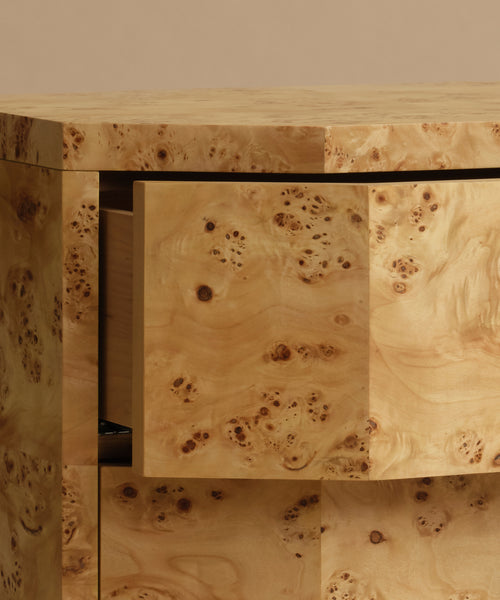 A close up of a burlwood nightstand with drawers, featuring natural burl wood grain and small dark metal knobs, set against a solid beige background.