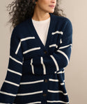 Wearing the Chloe Cardigan in Navy/Ivory Stripe with white horizontal stripes over a white shirt, a person stands against a neutral background. The V-neck cardigan has large front buttons and only the upper body is visible.