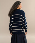 A woman with long curly hair, seen from behind, wears the Chloe Cardigan in Navy/Ivory Stripe and a dark skirt against a plain beige background.