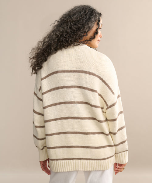 A person with long, curly hair faces away from the camera, modeling the Chloe Cardigan in ivory taupe stripe with white pants against a neutral backdrop—a stylish choice for warm-weather layering.