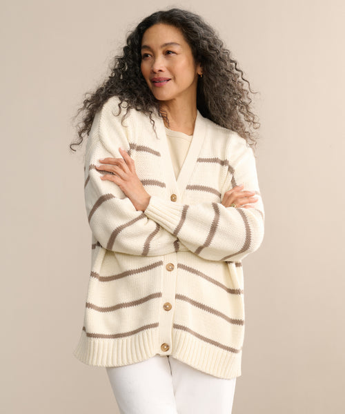 A woman with long curly hair wears the Chloe Cardigan in ivory taupe stripes over a beige top and white pants. With her arms crossed and smiling to the side, she shows how this cardigan brings effortless style to warm-weather looks.
