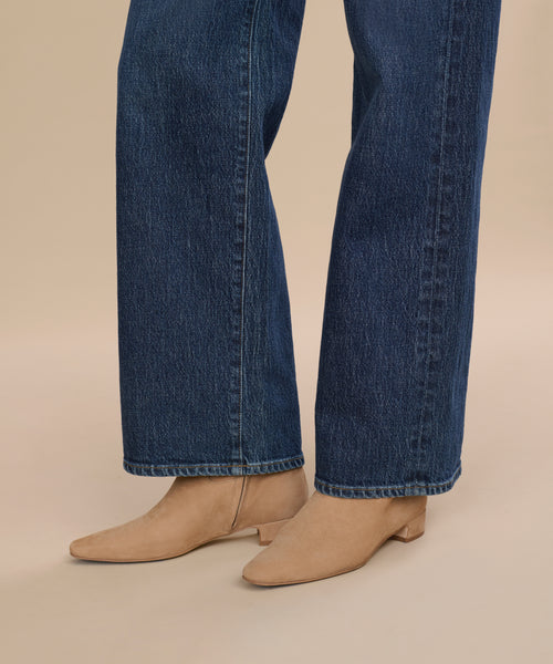 A person wearing wide-leg dark blue jeans and Suede Carmine Boot low-heeled taupe booties stands on a beige background.