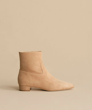 The Suede Carmine Boot features taupe suede, a low square heel, and a slightly pointed toe, shown against a matching beige background.