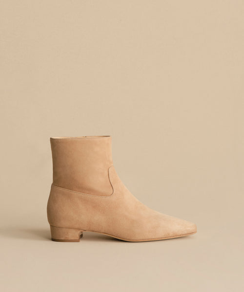 The Suede Carmine Boot features taupe suede, a low square heel, and a slightly pointed toe, shown against a matching beige background.