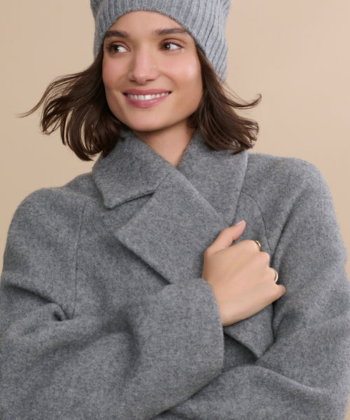A woman smiles and looks to the side against a beige background, wearing the Caroline Coat—an oversized grey wool coat—and a matching knit beanie.