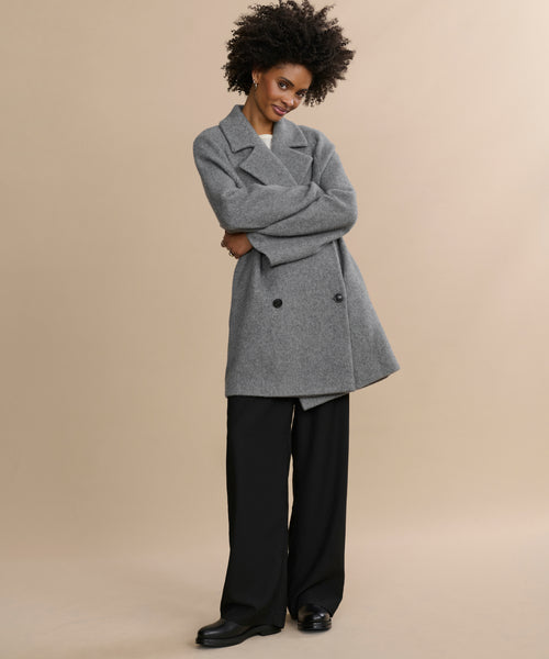 A woman with curly hair smiles while wearing the gray wool Caroline Coat, black wide-leg pants, and black shoes, standing against a plain beige background.