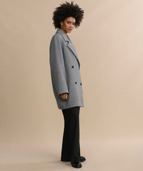 A woman with curly hair and a relaxed expression stands sideways against a beige background, wearing the Caroline Coat—an oversized gray double-breasted style—paired with black pants and black shoes.