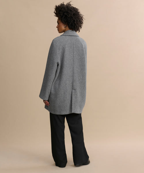 A person with curly hair stands facing away, wearing the oversized gray Caroline Coat and black pants against a neutral beige background.