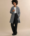 Against a beige background, a person with curly hair confidently poses, wearing the Caroline Coat—an oversized gray wool layer—over a white top and black wide-leg pants with black shoes, one hand holding the coat lapel.