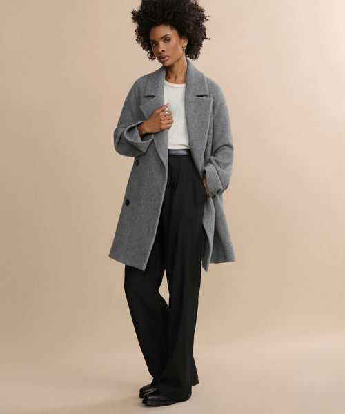 Against a beige background, a person with curly hair confidently poses, wearing the Caroline Coat—an oversized gray wool layer—over a white top and black wide-leg pants with black shoes, one hand holding the coat lapel.