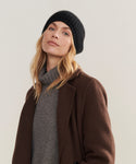 A person with wavy blonde hair wears the Cashmere Alpine Beanie—an essential black winter accessory—with a gray turtleneck and brown coat, standing against a neutral background.