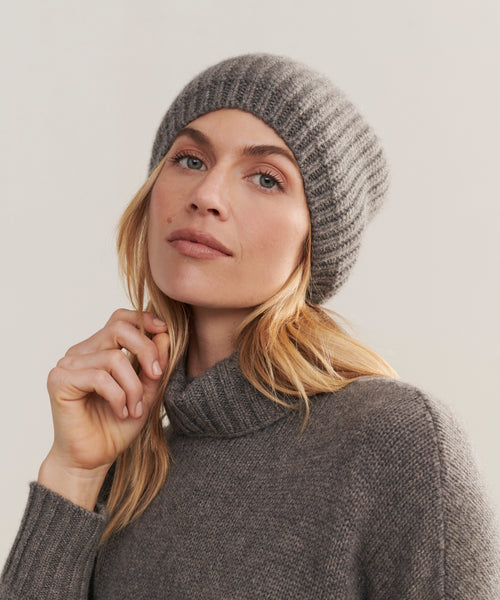 A woman wearing the Cashmere Alpine Beanie in ribbed gray and a gray turtleneck sweater gazes at the camera with a neutral expression, her hand resting on her chin against a light, plain background.