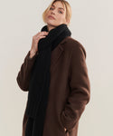 A person in a long brown coat wears the Cashmere Alpine Scarf in black, draping it stylishly over one hand while gazing slightly upward against a plain, light background.