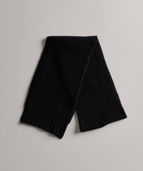 The Cashmere Alpine Scarf in black features a ribbed knit texture and is displayed flat on a light surface with its ends gently overlapping.