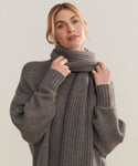 A person with light brown hair, wearing a cozy oversized gray sweater and the Cashmere Alpine Scarf, holds the chunky knit accessory close to their neck against a plain light background.