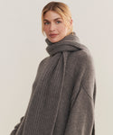 A woman with blonde hair in a low ponytail wears the Cashmere Alpine Scarf—a chunky gray knit winter accessory—along with a matching gray sweater, looking at the camera with a soft, neutral expression against a light background.