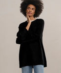 A woman with curly hair wears the Cashmere Amelia Crewneck in black over a white top and blue jeans, standing against a plain light background and thoughtfully touching her face with one hand.