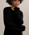 A woman with curly hair, wearing the Cashmere Amelia Crewneck in black, rests her face on her hand and gazes thoughtfully at the camera against a neutral background.