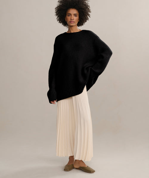 A woman with short curly hair and a calm expression wears the oversized black Cashmere Amelia Crewneck, a long pleated cream skirt, and brown flats against a neutral background.