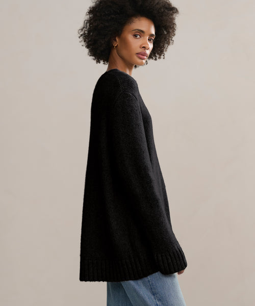 A woman with curly hair models the oversized black Cashmere Amelia Crewneck and light blue jeans, standing sideways against a plain light background as she gazes softly at the camera.